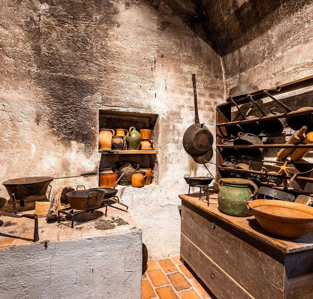 In der Ecke eines gemauerten Raumes stehen und hängen alte Küchengegenstände auf einer Ablage und einem alten Holzschrank mit Regal.