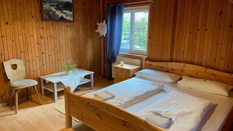 Holzvert&auml;feltes Doppelzimmer mit Bett, Tisch, Stuhl und Fenster.