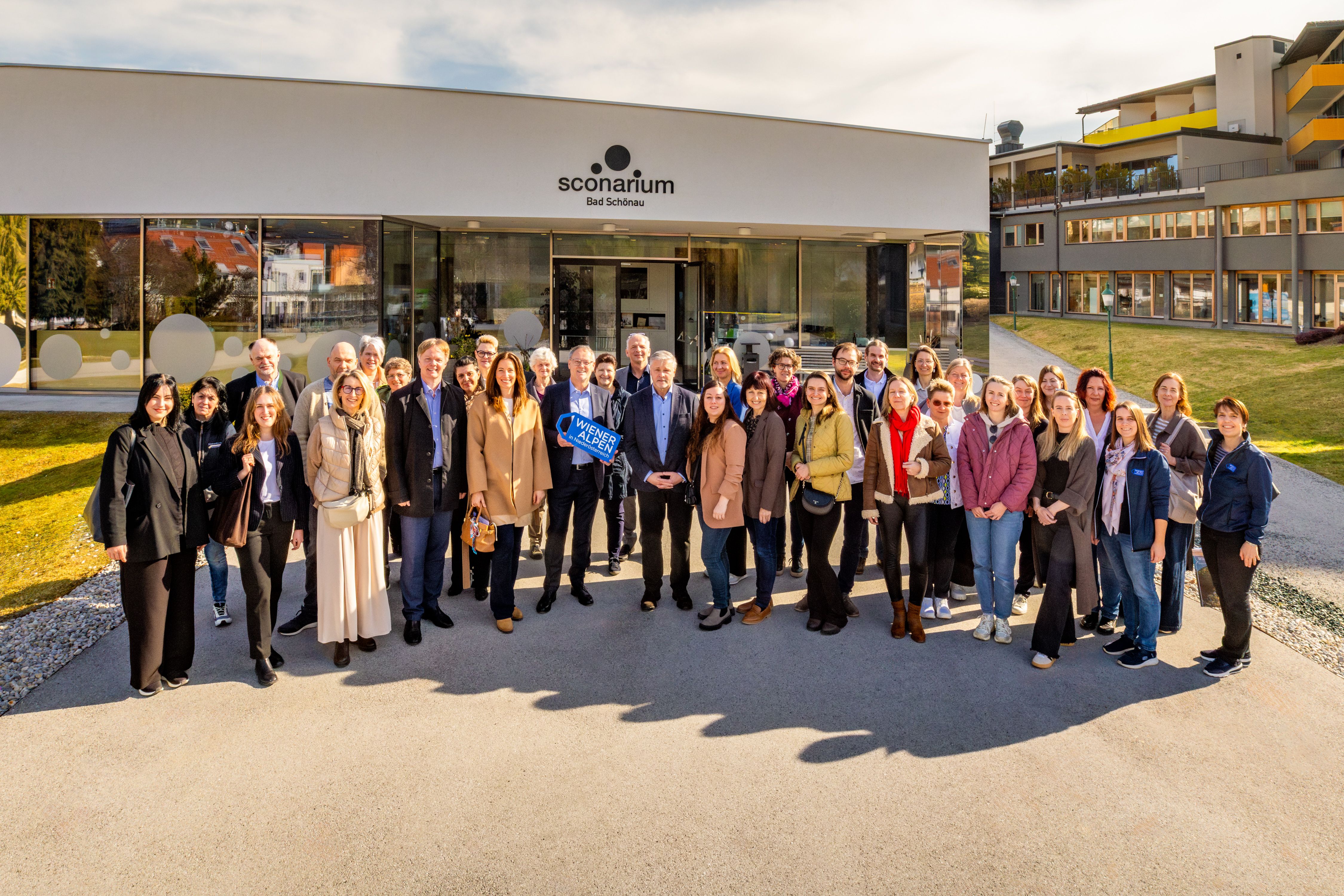 Rund 40 Personen stehen bei Sonnenschein am Vorplatz eines modernen Gebäudes (dem Kulturhaus) mit Glasfront und der Aufschrift "Sconarium Bad Schönau". Der Bürgermeister der Gemeinde Bad Schönau hält ein Logoschild in den Händen.