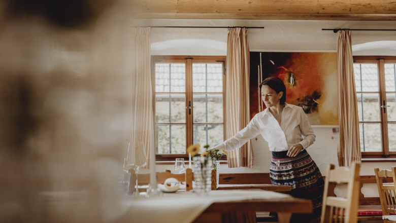 A woman in traditional dress sets up a table in a cozy room with wooden furniture and large windows.