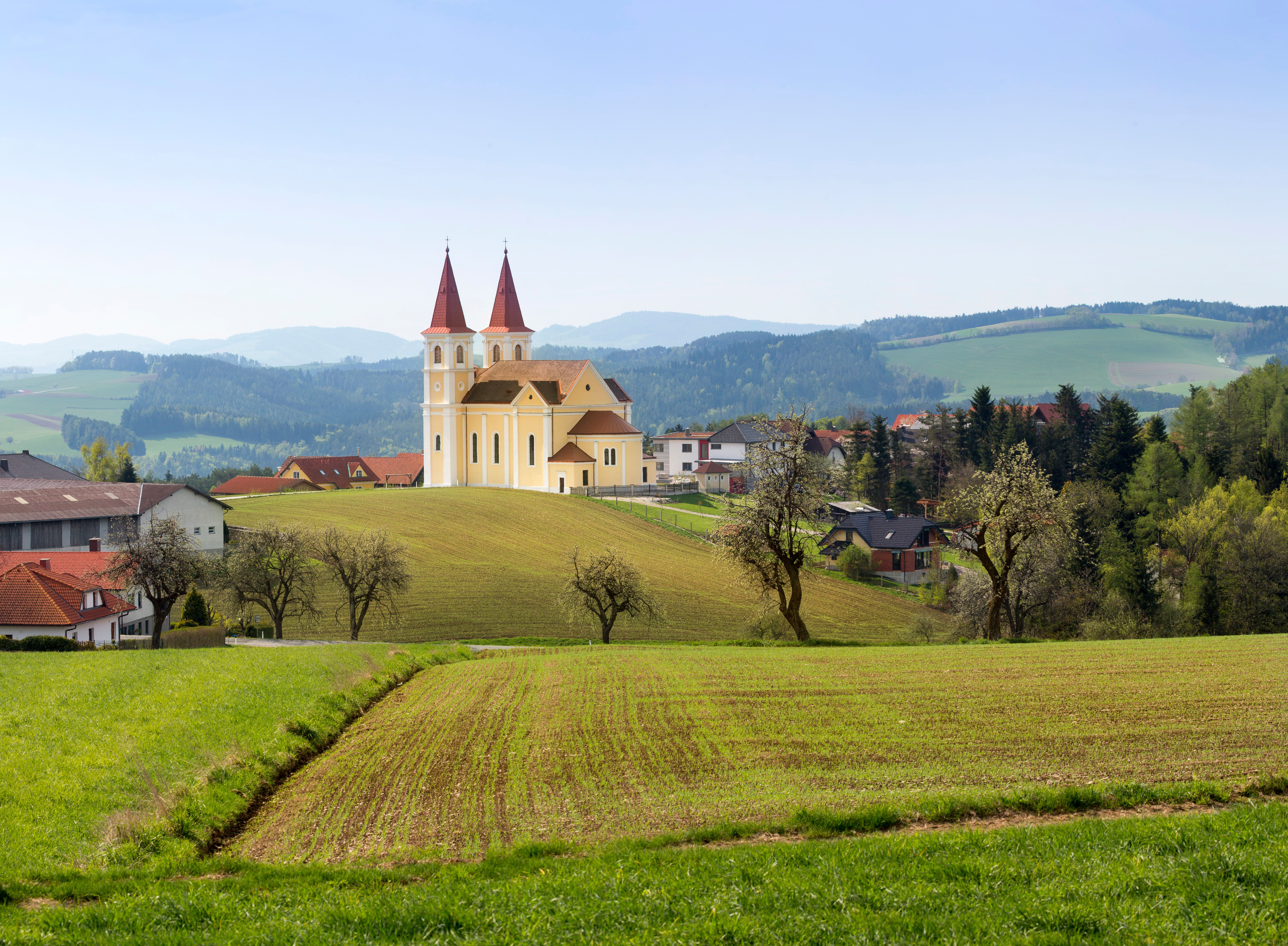 Eine Kirche mit zwei Türmen steht auf einem Hügel in einer ländlichen Landschaft mit Feldern und Bäumen.
