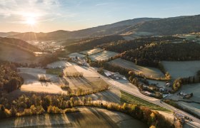 Luftaufnahme einer ländlichen Landschaft bei Sonnenaufgang mit Feldern, Wäldern und vereinzelten Gebäuden.