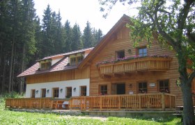 A rustic wooden mountain hut with balcony and terrace, surrounded by trees.