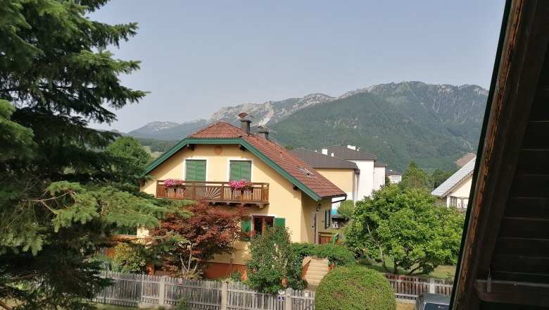 Blick von einem Balkon auf ein gelbes Haus mit rotem Dach, umgeben von B&auml;umen und Bergen im Hintergrund.