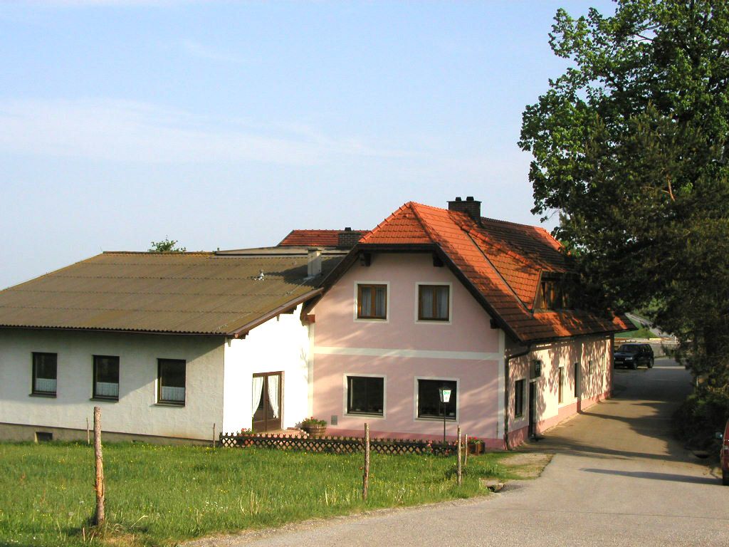 Ein traditionelles Gasthaus mit rosa und weißen Wänden und einem roten Ziegeldach, umgeben von grüner Landschaft.