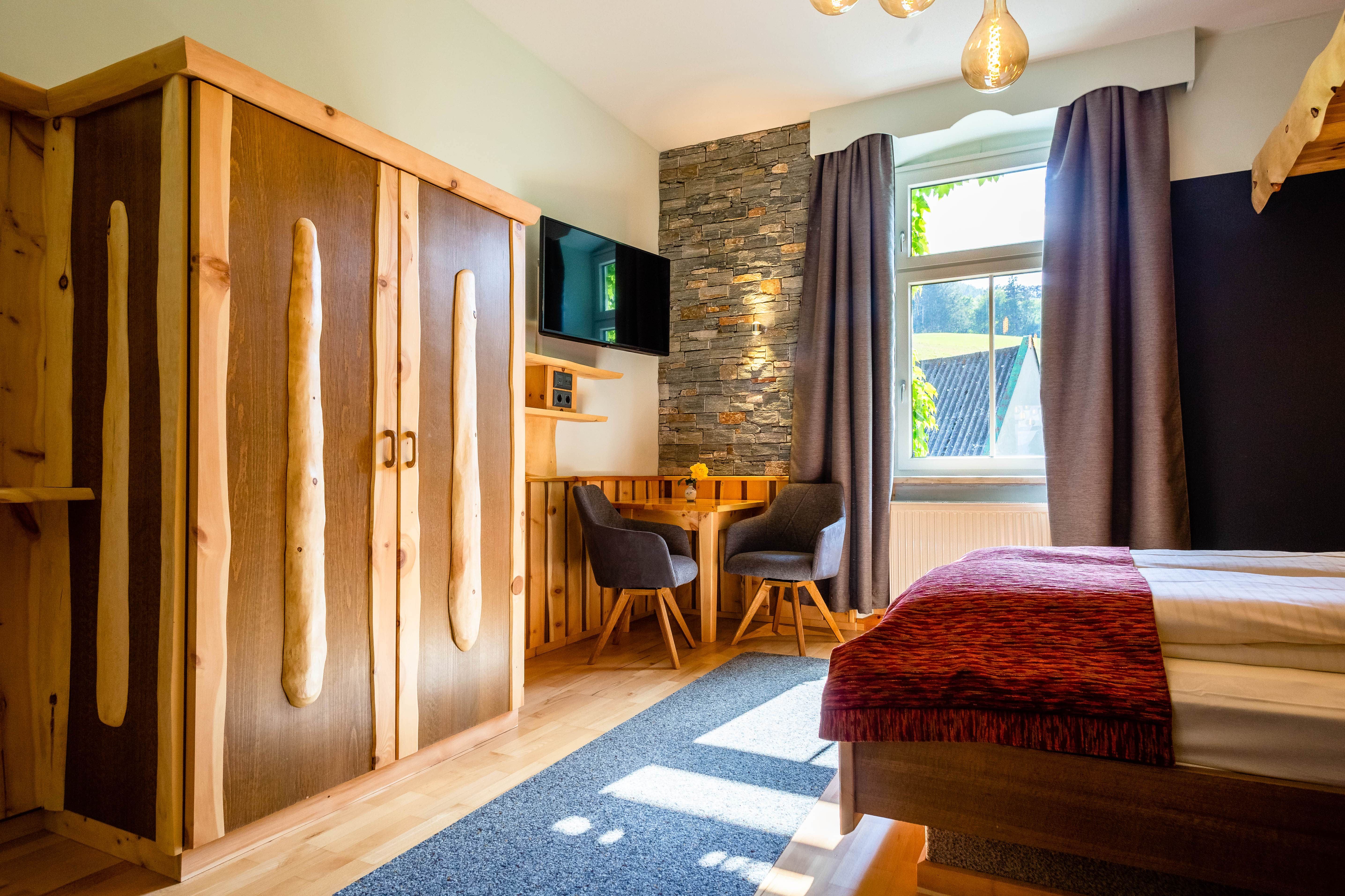 Gemütliches Zimmer mit Holzmöbeln, Steinwand, Tisch und Stühlen, Fernseher und Fenster mit Vorhängen.