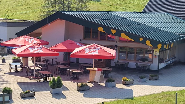 Außenansicht eines Restaurants mit roten Sonnenschirmen und Sitzplätzen im Freien.