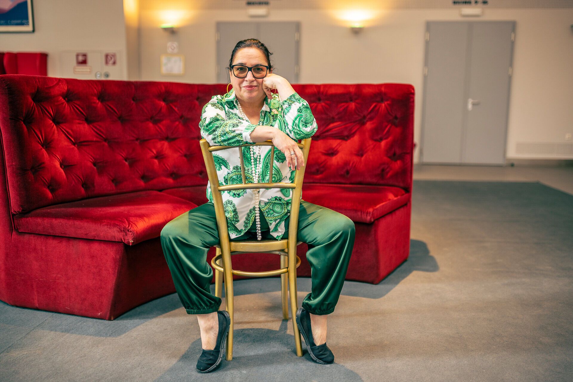 Frau mit Brille sitzt lässig auf einem Stuhl vor einer roten Samtbank in einem modernen Innenraum.