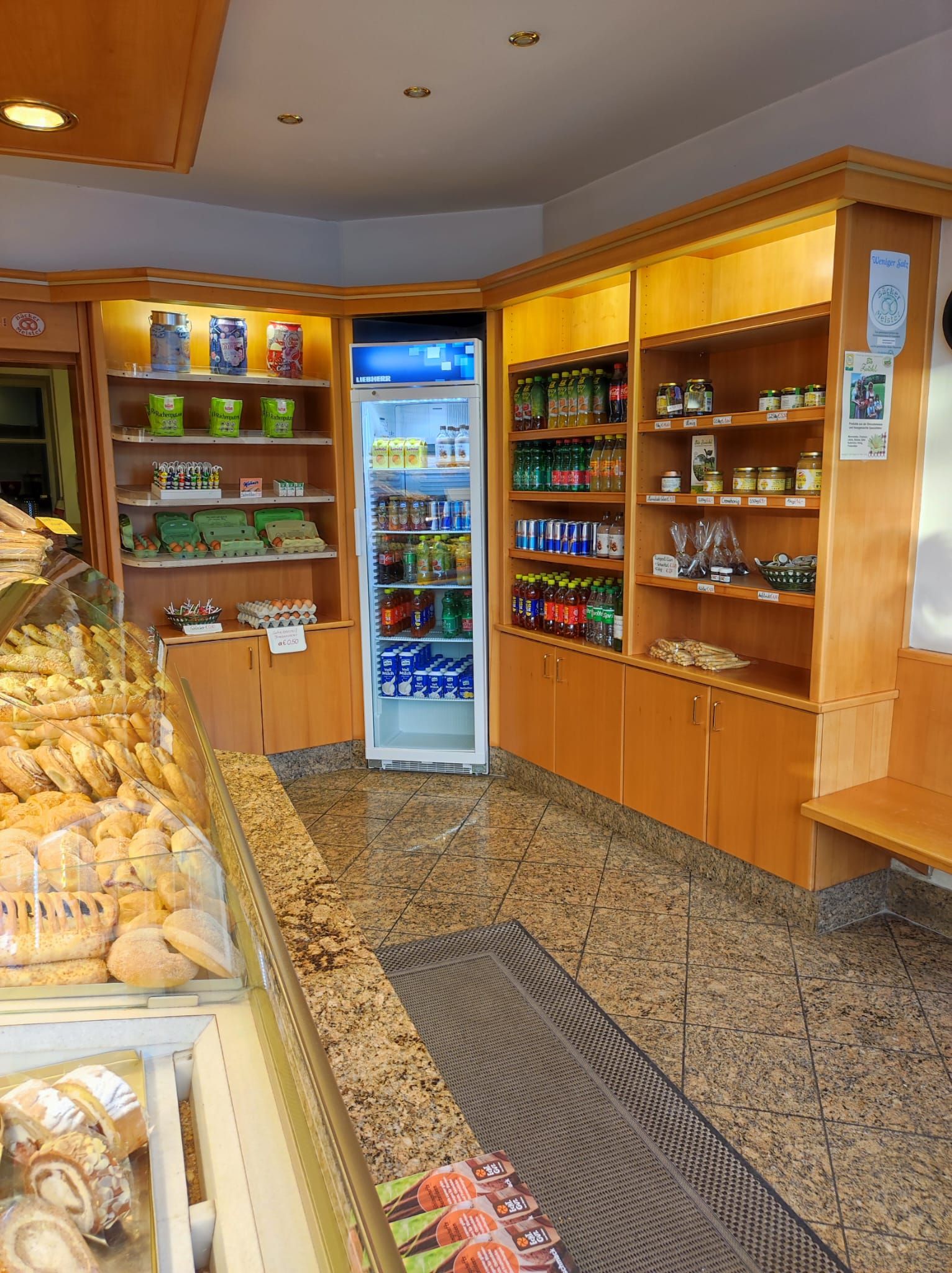 Innenraum von Cafe Dietmayer in Ternitz mit Backwarenvitrine, Getränkekühlschrank und Holzregalen voller Snacks und Getränke.