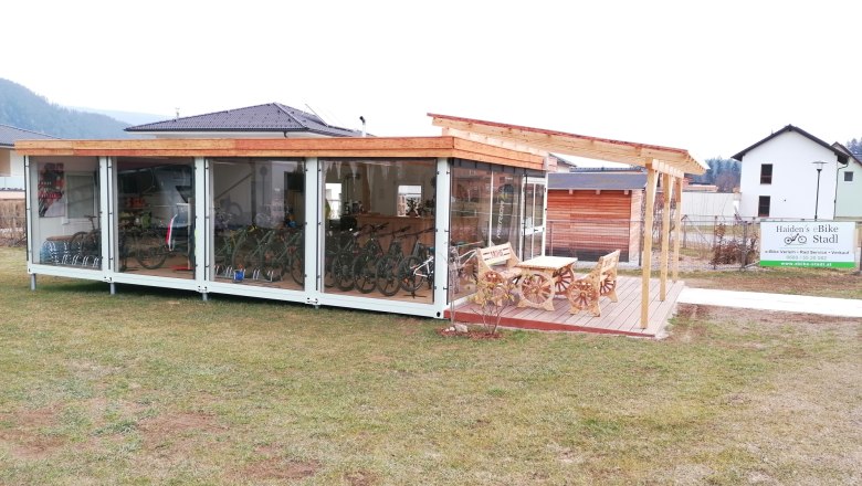 Außenansicht eines modernen Fahrradverleihs mit Glasfront und Holzdach, umgeben von Rasen und einem Schild mit der Aufschrift 'Haiden's eBike Stadl'.