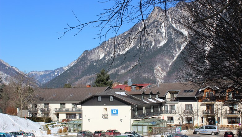 Außenansicht eines Gebäudes vor einer Bergkulisse mit schneebedeckten Gipfeln und blauem Himmel.