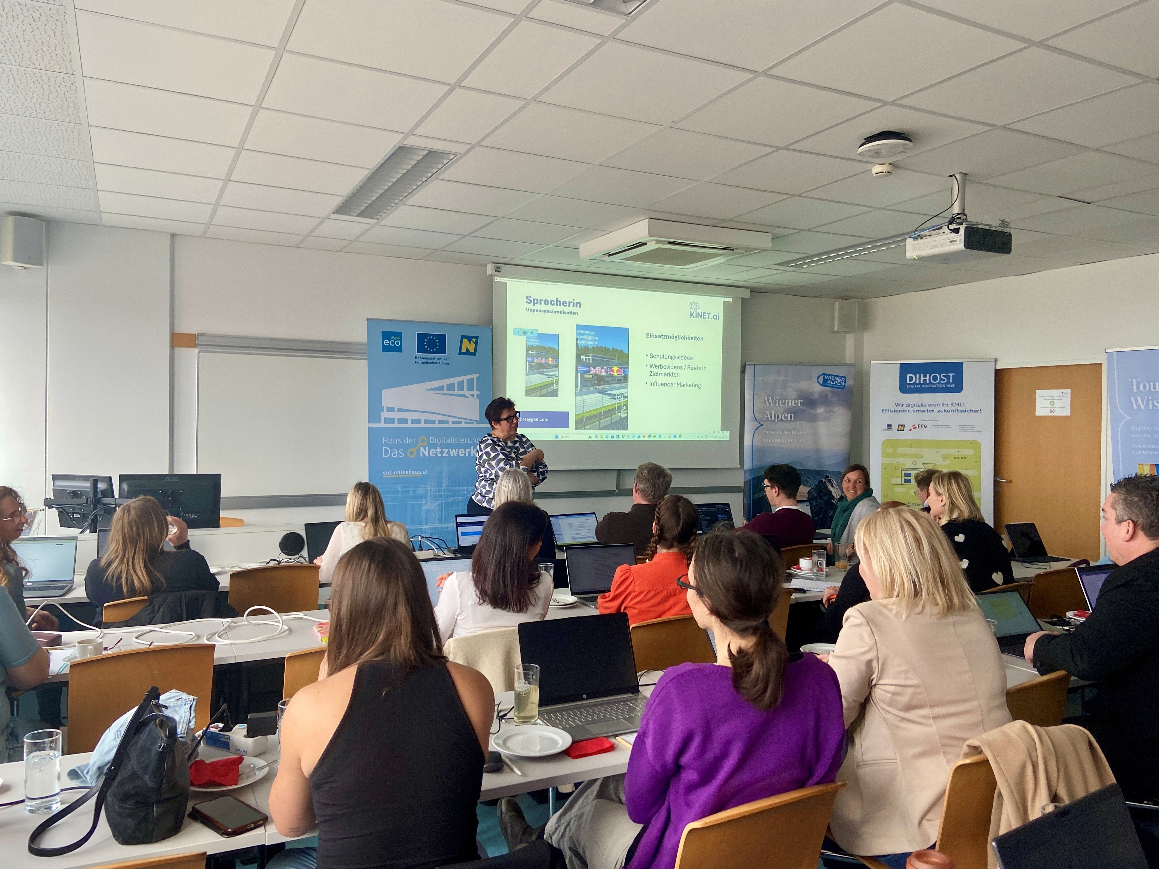 In einem Schulungsraum sitzen mehrere Teilnehmer eines Workshops mit dem Rücken zur Kamera in mehreren Reihen. Davor steht eine Person vor mehreren Roll-Ups und einer Leinwand.