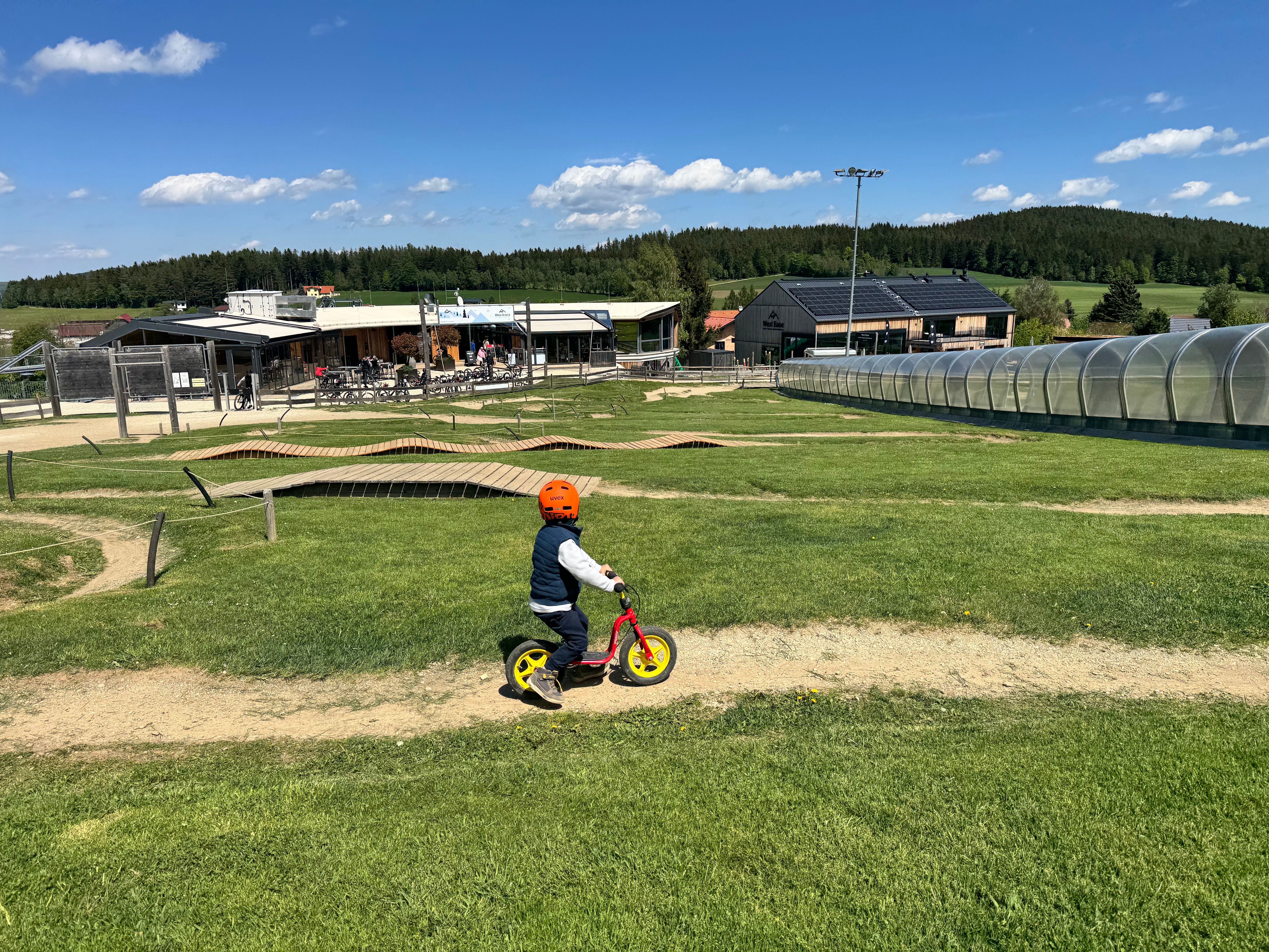 Ein Kind mit Helm fährt auf einem Laufrad über einen grasbewachsenen Hügel. Im Hintergrund sind Gebäude und ein überdachter Gehweg zu sehen.
