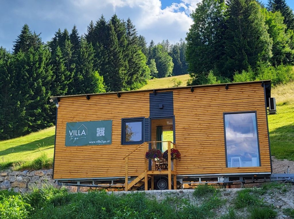 Kleine Holzhütte auf Rädern vor einem bewaldeten Hügel.