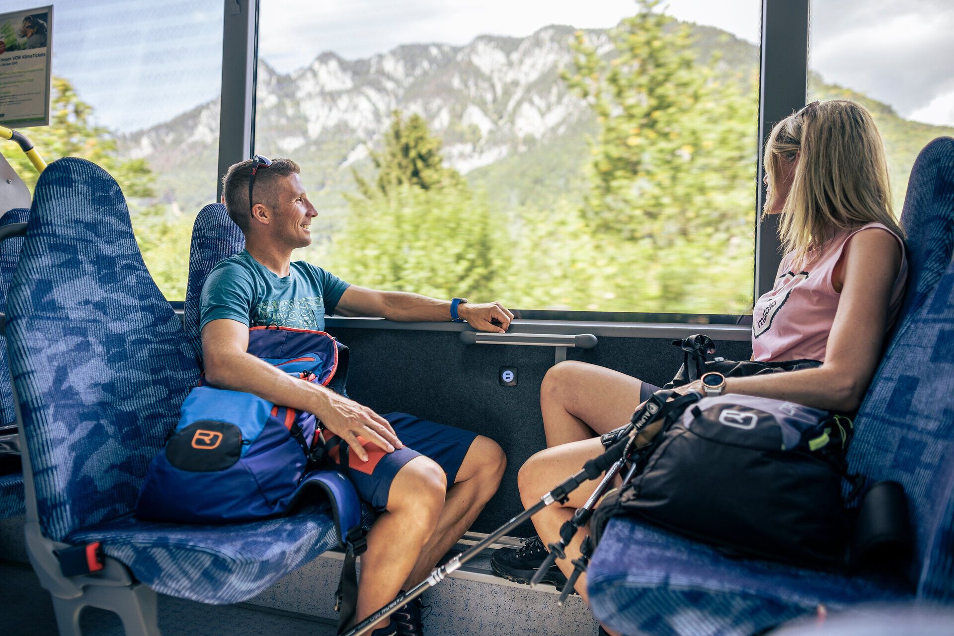 Zwei Fahrgäste sitzen im Bus und genießen den Ausblick auf eine alpine Landschaft.