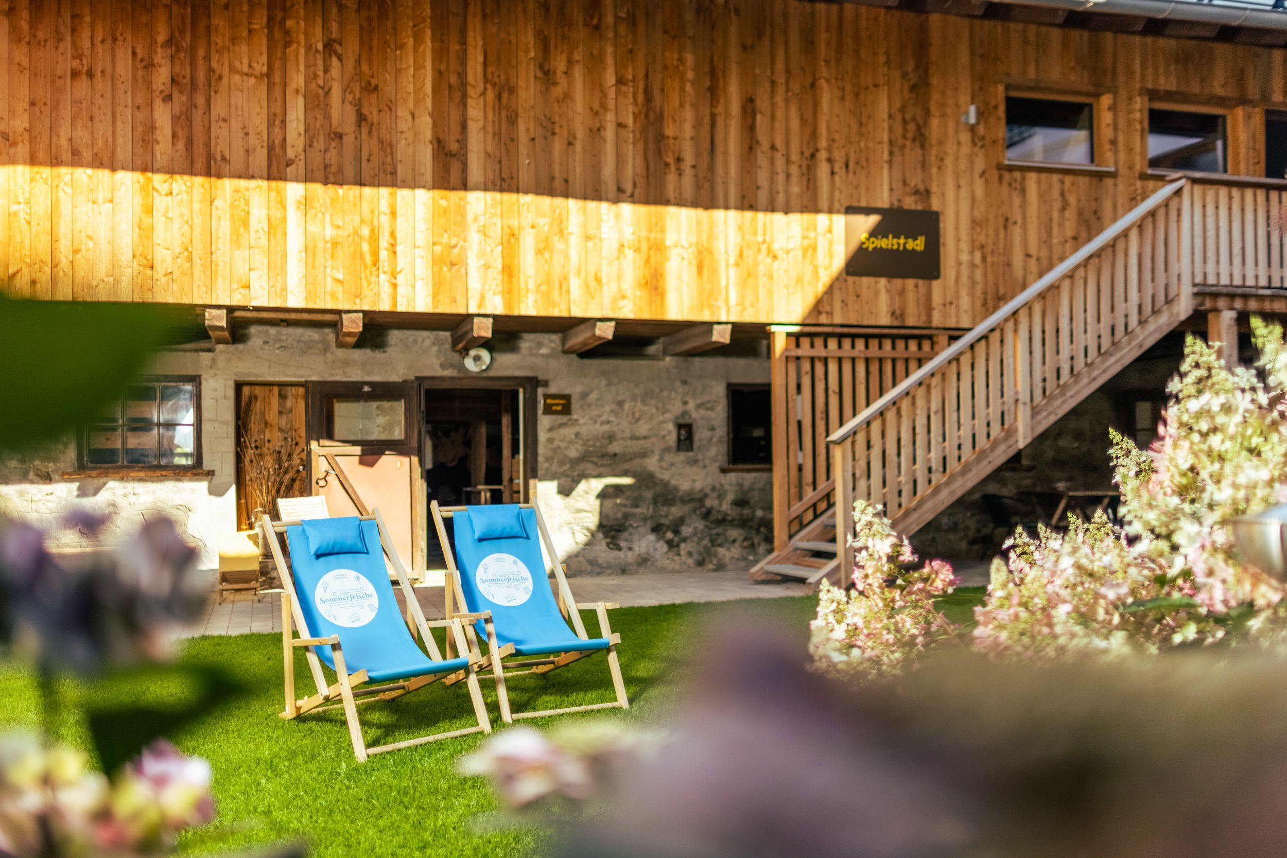 Zwei blaue Liegestühle auf einer Wiese vor einem rustikalen Holzgebäude.