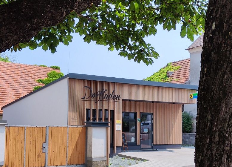 Ein moderner Dorfladen mit Holzfassade und Schildern, umgeben von Bäumen.