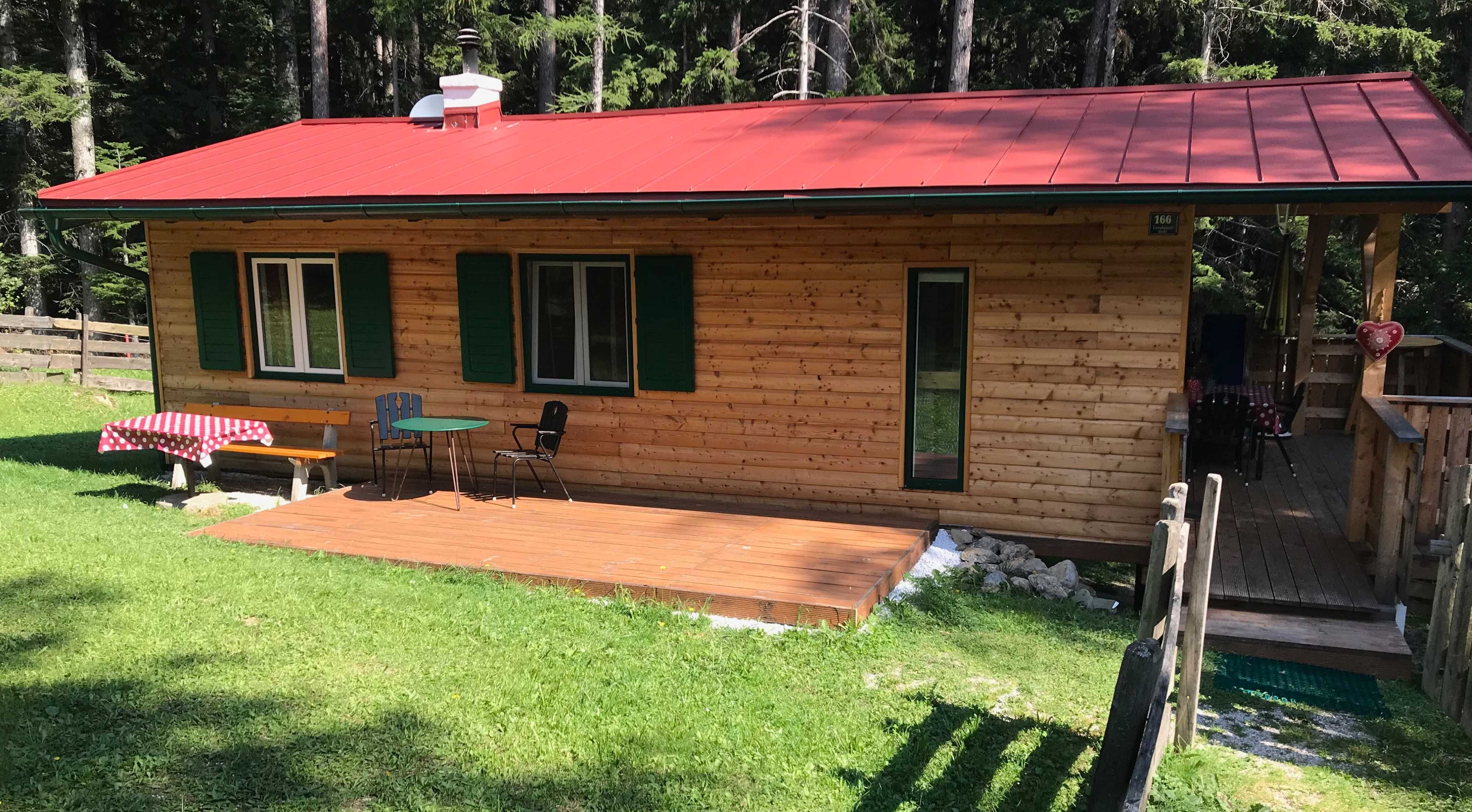 Ein kleines Holzferienhaus mit rotem Dach, Terrasse und Gartenmöbeln, umgeben von Wald.
