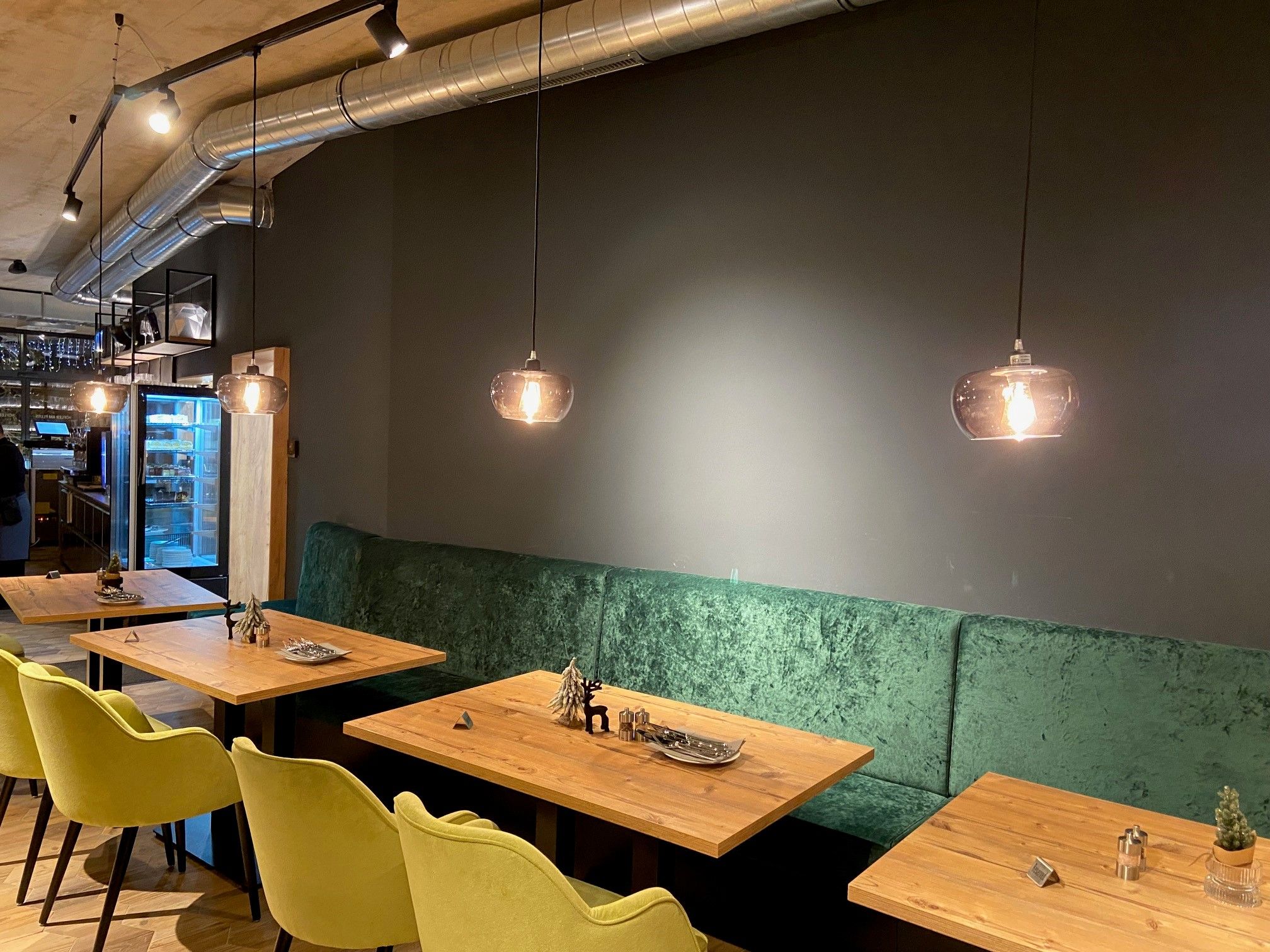 Innenansicht eines modernen Restaurants mit Holztischen, grünen gepolsterten Bänken und hängenden Lampen.