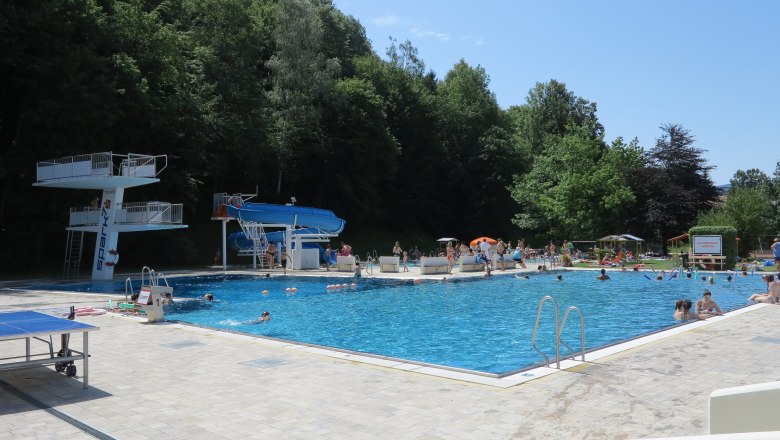 Ein Freibad mit Sprungturm, Rutsche und vielen Badegästen an einem sonnigen Tag.