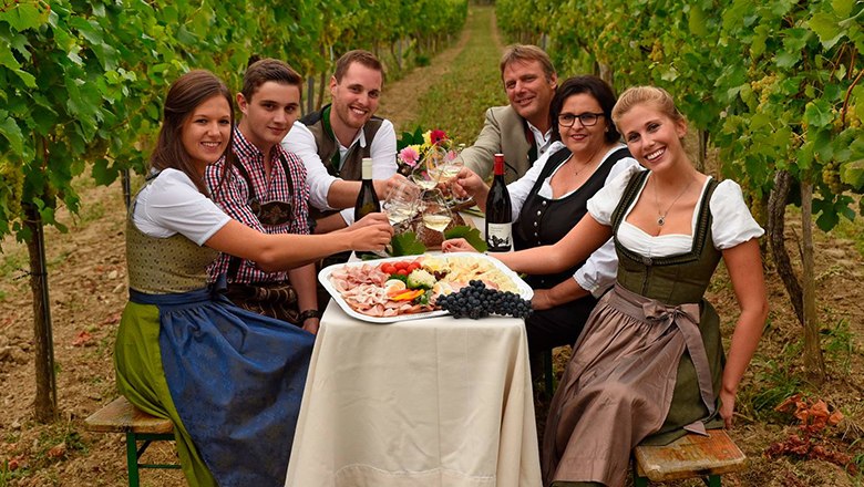 A family sits at a table with wine, platters of cold meats and glasses of wine between vines.