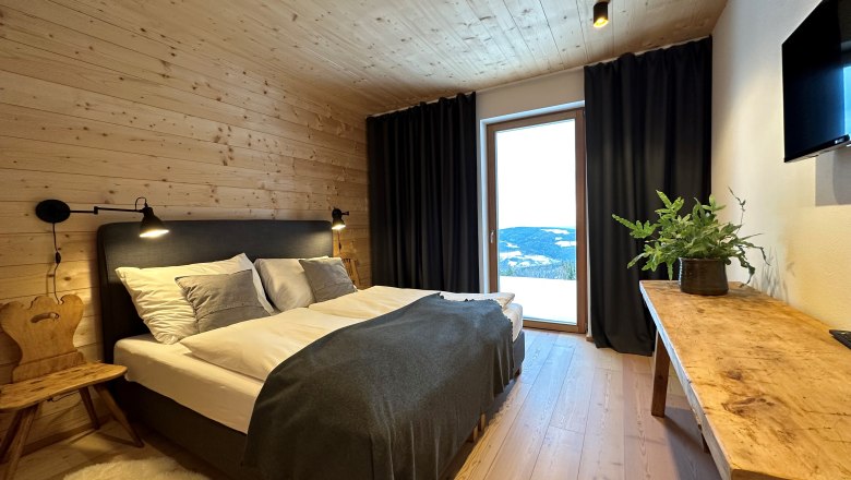 Gemütliches Schlafzimmer mit Holzwänden, Doppelbett, Holzstuhl, Tisch mit Pflanze und großem Fenster mit Vorhängen.