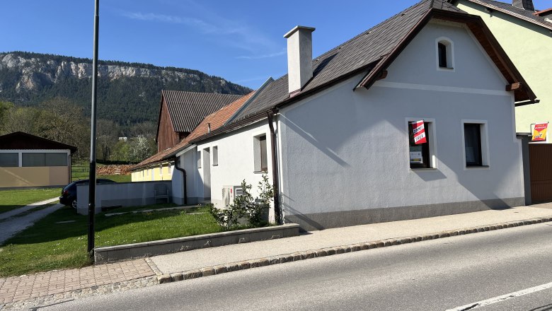 A white house with a red roof and a "For Rent" sign in front of a mountain backdrop.