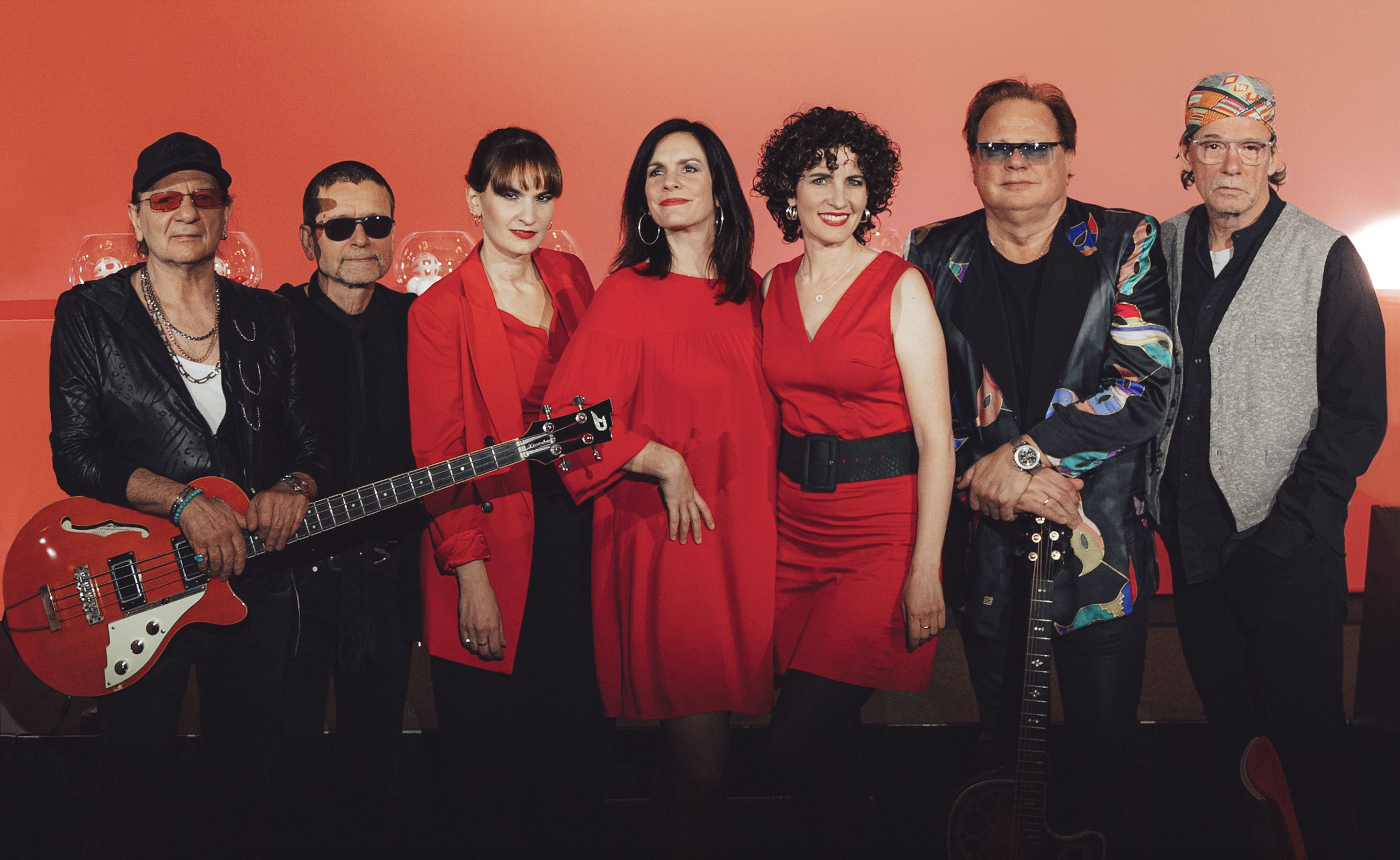 drei Damen in roter Kleidung und vier Männer in schwarzer Kleidung vor rotem Hintergrund mit Musikinstrumenten