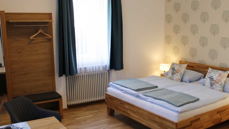 Gem&uuml;tliches Doppelzimmer mit Bett, Kleiderschrank, Tisch, Stuhl und Fenster mit blauen Vorh&auml;ngen.