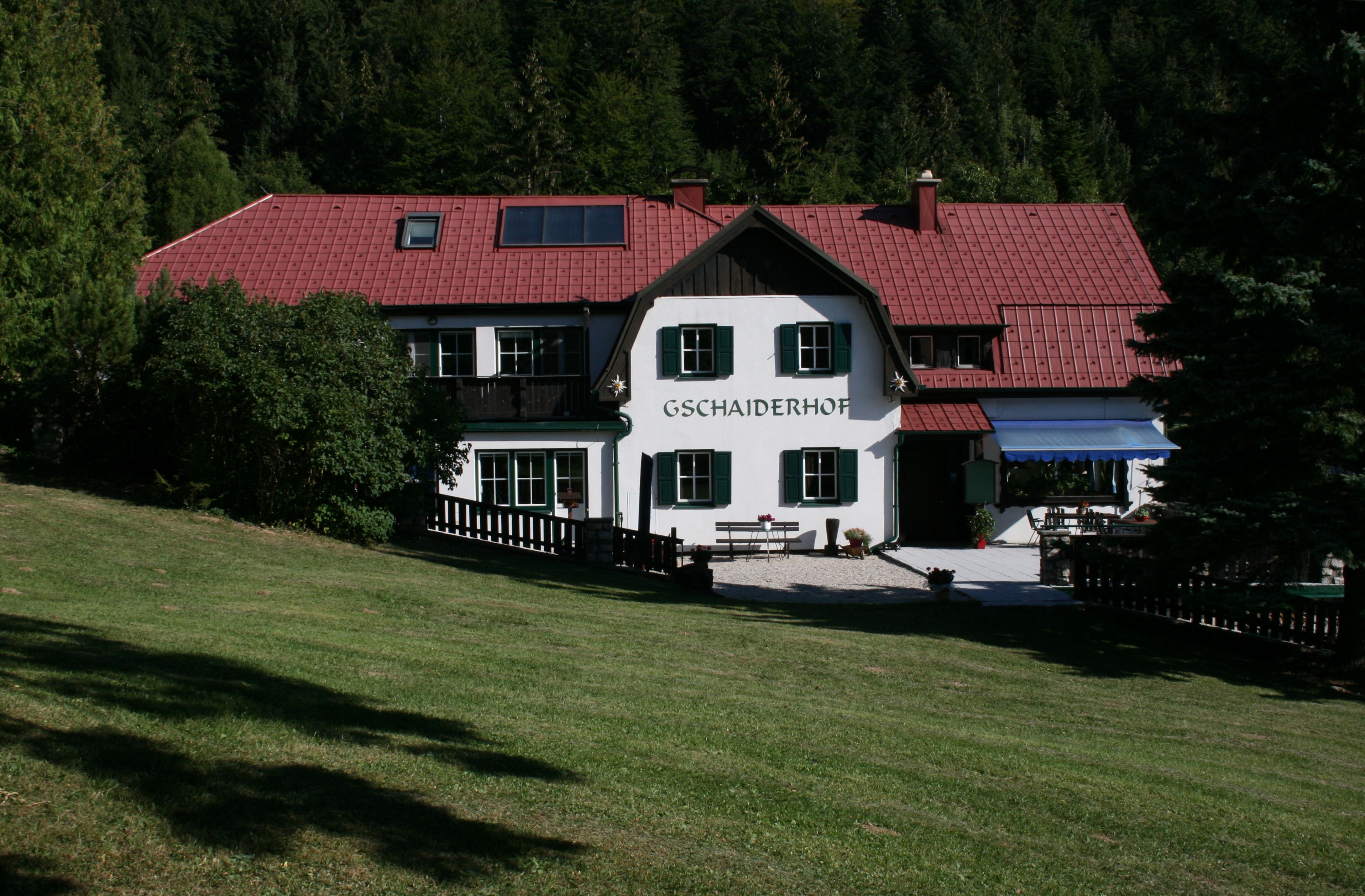 Ein traditionelles Gebäude mit rotem Dach und grünen Fensterläden, umgeben von Bäumen und Wiese, mit dem Schriftzug 'Gschaiderhof'.