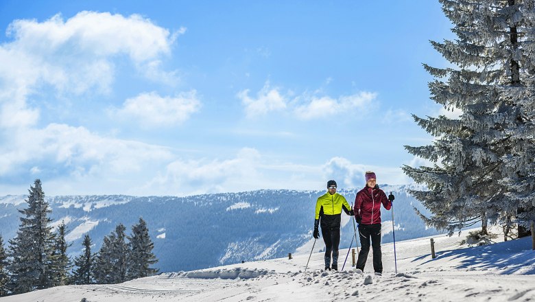 Zwei Langläufer auf einer schneebedeckten Loipe in einer winterlichen Berglandschaft.