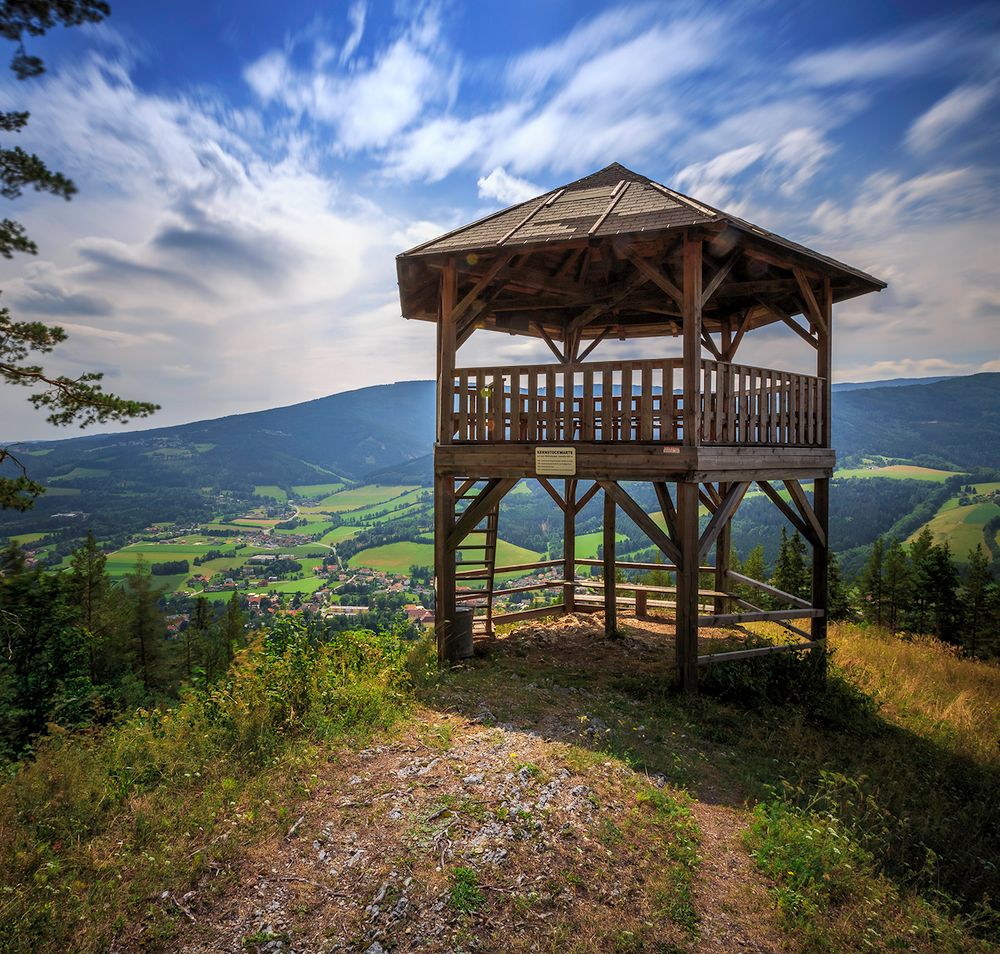 Holzaussichtsturm mit Blick auf grüne Täler und Berge unter blauem Himmel.