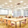 A bright seminar room with a U-shaped table arrangement, screen, flipchart and drinks table.