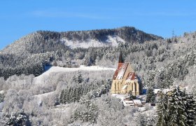 Winterlandschaft mit Kirche vor bewaldetem Hügel.