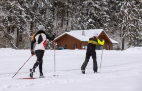 Zwei Langläufer auf einer verschneiten Loipe vor einer Holzhütte im Wald.