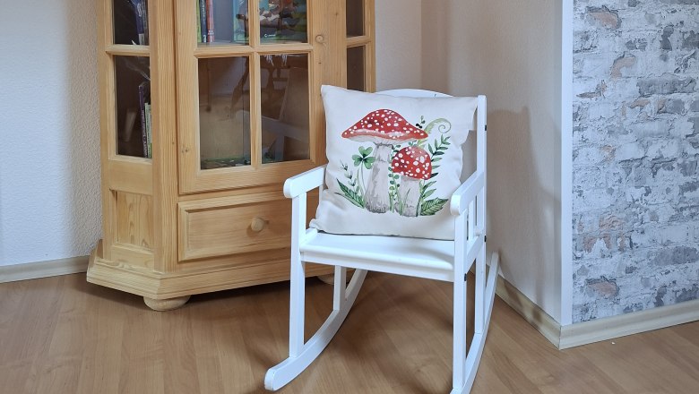 Ein Holzschrank mit Büchern und ein weißer Schaukelstuhl mit einem Kissen, das Pilze zeigt, stehen in einem Raum mit Holzboden und gemusterter Tapete.