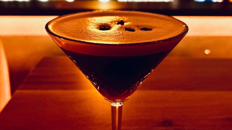 Ein Cocktailglas mit dunklem Getränk auf einem Holztisch, im Hintergrund eine unscharfe Bar.