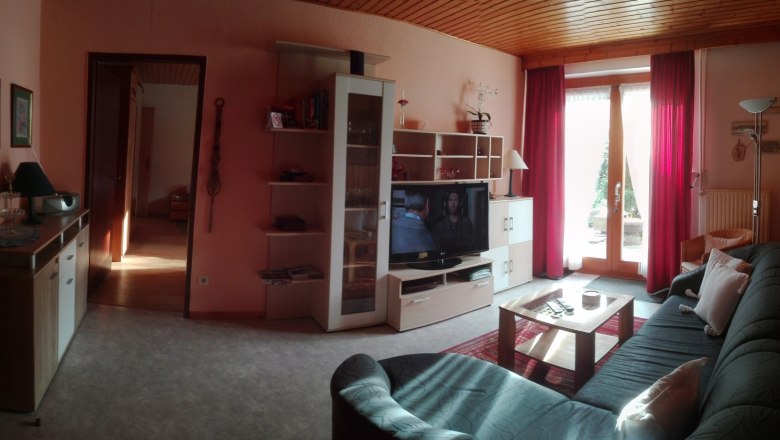 Gemütliches Wohnzimmer mit Sofa, Fernseher, Regal und Glastür mit roten Vorhängen.