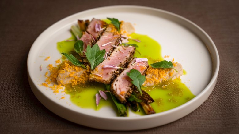 Ein kunstvoll angerichtetes Gericht mit Fleisch, grüner Sauce und essbaren Blüten auf einem weißen Teller.