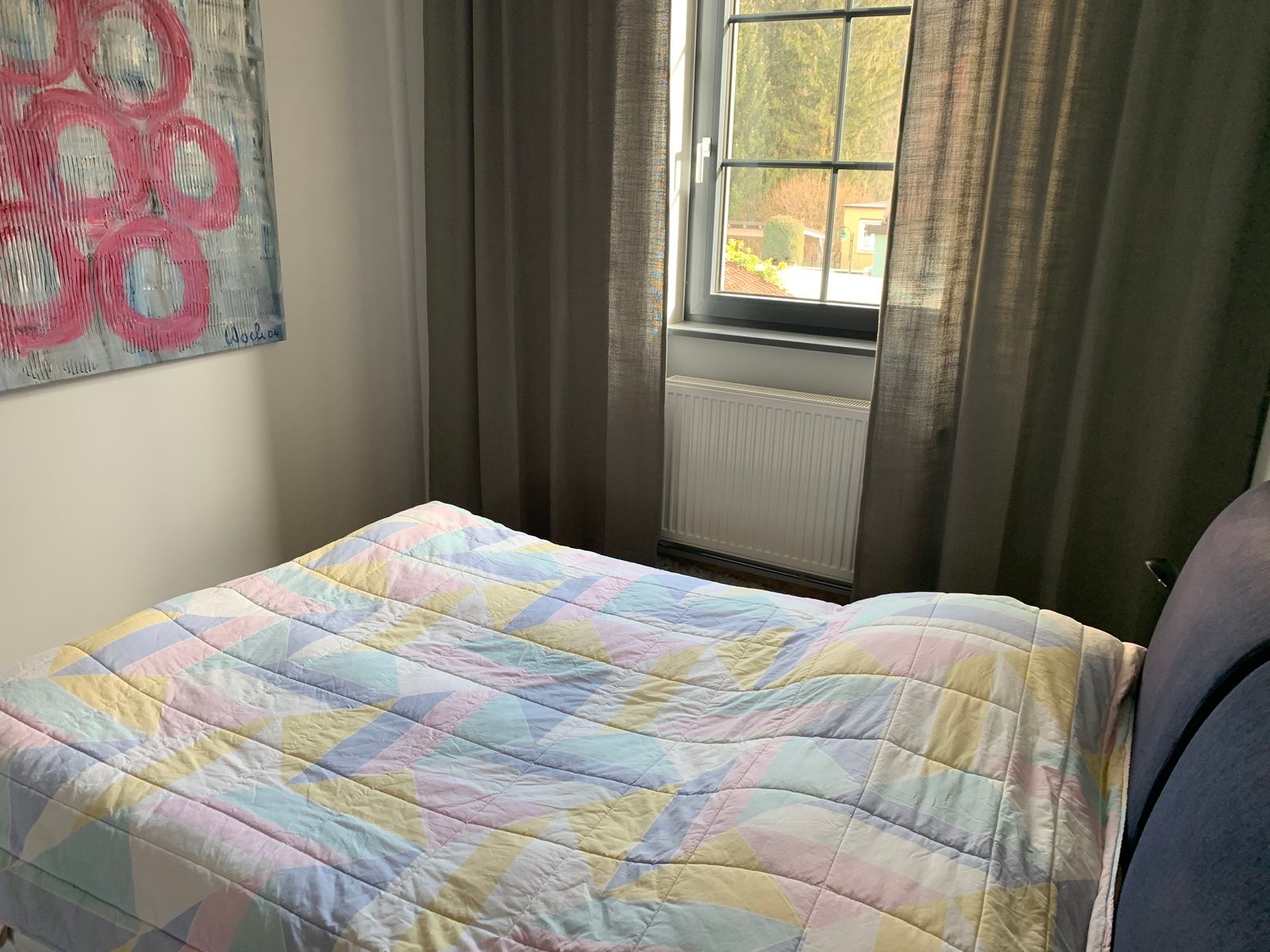 Schlafzimmer mit Bett, bunter Decke, Fenster und Kunstwerk an der Wand.