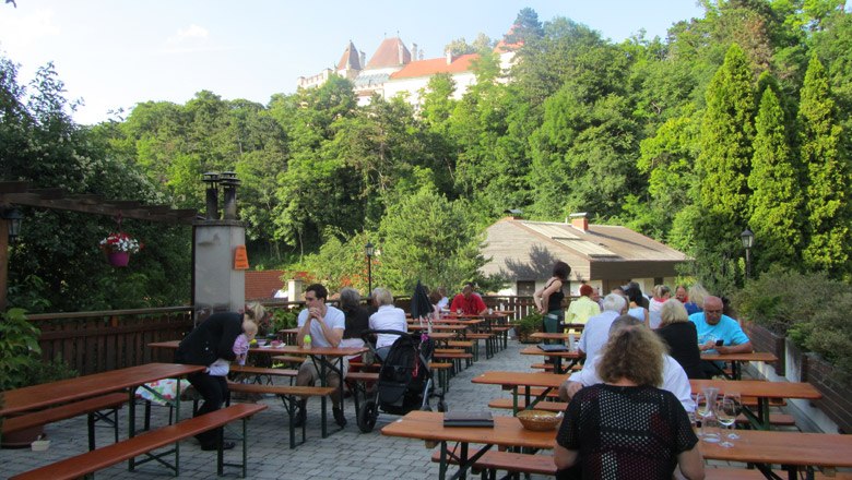 People sit at wooden tables outside with a view of a castle in the background.