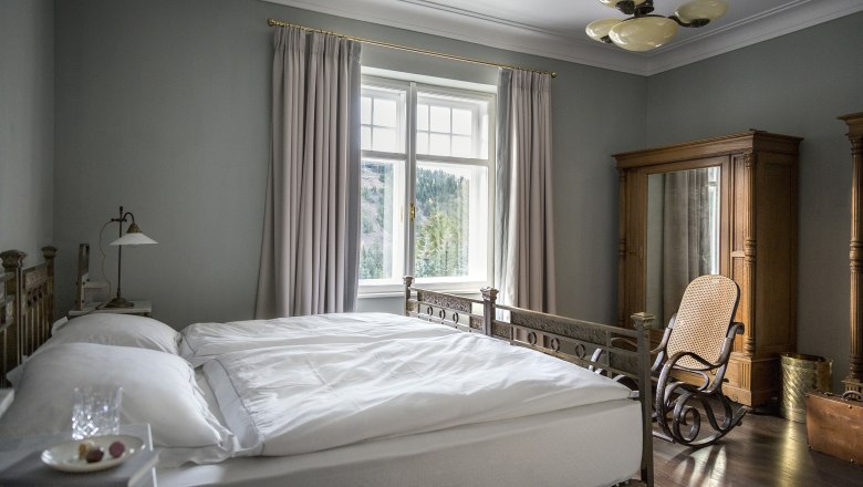 Elegantes Schlafzimmer mit antikem Bett, Schaukelstuhl und gro&szlig;em Fenster mit Vorh&auml;ngen.