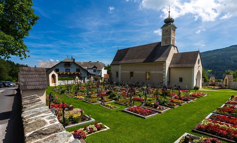 Pfarrkirche St. Peter am Neuwald mit gepflegtem Friedhof und blühenden Blumenbeeten, umgeben von grüner Landschaft und blauem Himmel.