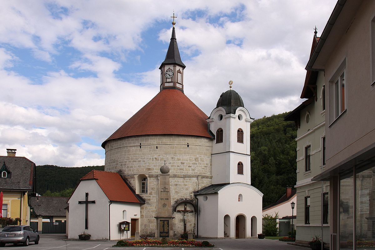 Pfarrkirche Scheiblingkirchen mit rundem Turm und rotem Dach, umgeben von Gebäuden und bewölktem Himmel.