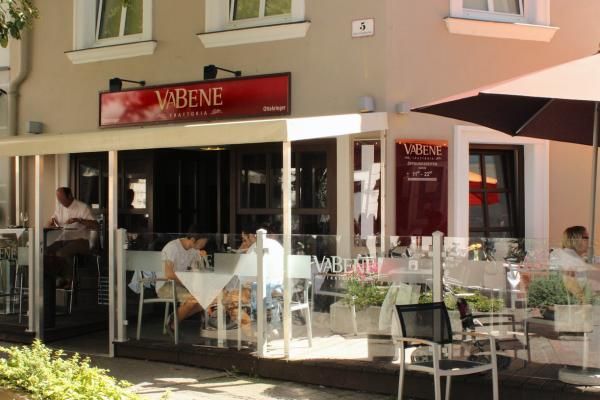 Außenansicht eines Cafés namens Va Bene mit Schanigarten und Gästen an Tischen.