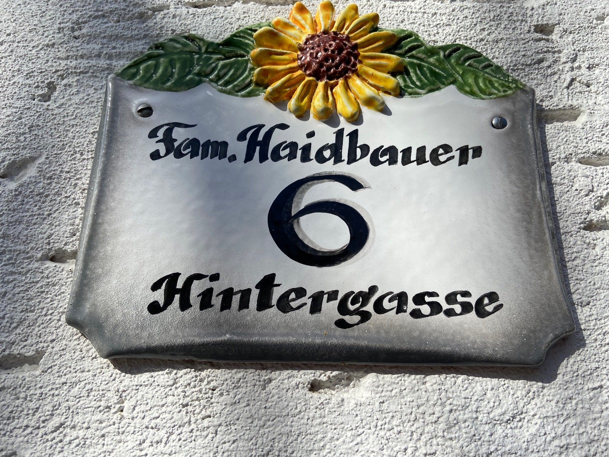 Hausnummernschild mit Sonnenblume und der Aufschrift 'Fam. Haidbauer 6 Hintergasse'.