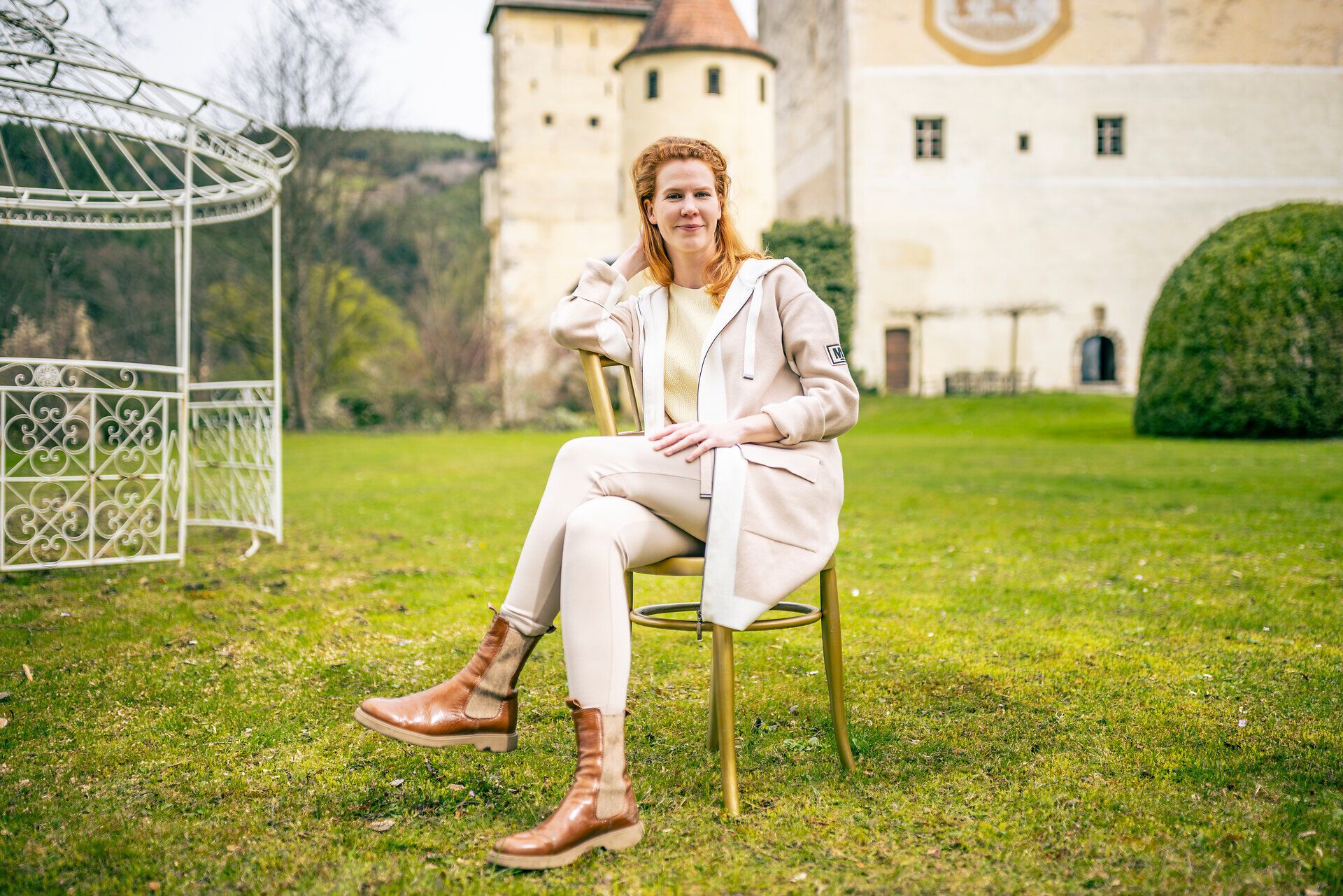 Frau sitzt auf einer Wiese vor einem historischen Schlossgebäude und blickt lächelnd in die Kamera.