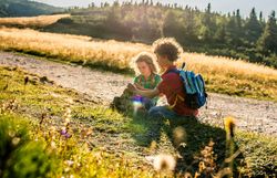 Zwei Kinder sitzen auf einer Wiese und erkunden die Natur, während die Sonne sanft über den Horizont strahlt. Umgeben von üppigem Grün und dem sanften Rauschen des Windes, erleben sie die Freude und Unbeschwertheit eines Sommertages in den Bergen.
