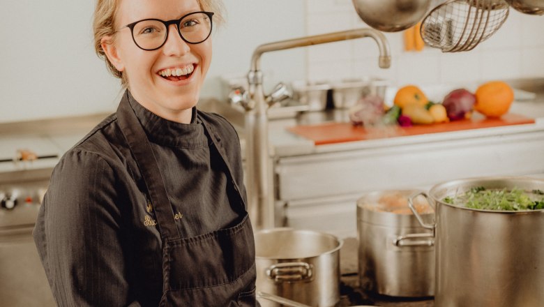 Kuchař v čern&eacute; uniformě stoj&iacute; s &uacute;směvem v profesion&aacute;ln&iacute; kuchyni a smaž&iacute; na p&aacute;nvi kus masa.