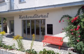 Exterior view of a building with the inscription 'Kurkonditorei', red bench and blooming roses.